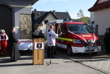 na placu przed remizą OSP w Stadnikach stoi lekki samochód ratowniczo-gaśniczy, z lewej strony samochodu stoi ksiądz i święci nowy nabytek, w centrum zdjęcia widać mównicę i mikrofon, z tyłu za mównicą stoi stół z białym obrusem na którym ustawione są statuetki