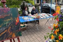 Zdjęcie na którym są dwie osoby i stół z rzeczami wystawionymi na sprzedaż.