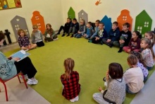 dzieci siedzą na dywanie tworząc okrąg i słuchają jak starsza koleżamka czyta im książkę