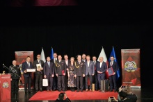 Grupa kilkunastu osób, w tym strażacy w mundurach i mężczyźni w garniturach oraz jedna kobieta w eleganckiej sukni, stoi na scenie na czerwonym dywanie podczas uroczystości. W tle widoczne są flagi Polski i Unii Europejskiej oraz banery z logo Państwowej Straży Pożarnej i adresem strony internetowej. Część uczestników trzyma dyplomy i upominki, a przed sceną dwie osoby fotografują wydarzenie.