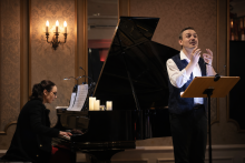 Mężczyzna śpiewa przy pulpicie nutowym podczas kameralnego koncertu, gestykulując rękami. Obok niego kobieta gra na fortepianie. Wnętrze jest eleganckie, o ciepłym oświetleniu – na ścianie widać ozdobny kinkiet, a na fortepianie stoją zapalone świece. Atmosfera sugeruje występ muzyki klasycznej lub recital.