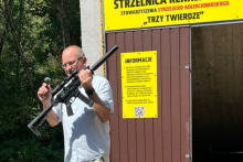 mężczyzna trzymający w lewej ręce broń w prawej mikrofon, w tle wejście do rekreacyjnej strzelnicy