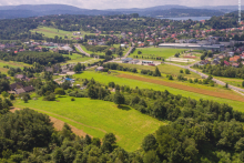 Widok z lotu ptaka na rozległy, zielony teren gminy – pola uprawne, łąki i zadrzewienia przeplatają się z zabudową jednorodzinną oraz siecią dróg lokalnych; w tle widoczne pagórkowate krajobrazy i zbiornik wodny.