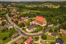 Widok z lotu ptaka na malowniczą miejscowość z zabudową jednorodzinną, otoczoną zielonymi wzgórzami i polami. Na pierwszym planie widoczny kościół oraz większy budynek z czerwonym dachem, przy których krzyżują się drogi. Wokół liczne domy, drzewa i kręta szosa prowadząca przez wieś.