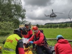 Mężczyźni z grupy GOPR podczas szkolenia na zielonej łące. Nad nimi unosi się helikopter