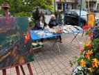Zdjęcie na którym są dwie osoby i stół z rzeczami wystawionymi na sprzedaż.