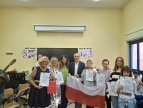 Grupa uczniów i nauczycieli prezentuje certyfikaty Erasmus+ oraz polską flagę w sali szkolnej.