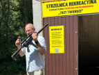mężczyzna trzymający w lewej ręce broń w prawej mikrofon, w tle wejście do rekreacyjnej strzelnicy