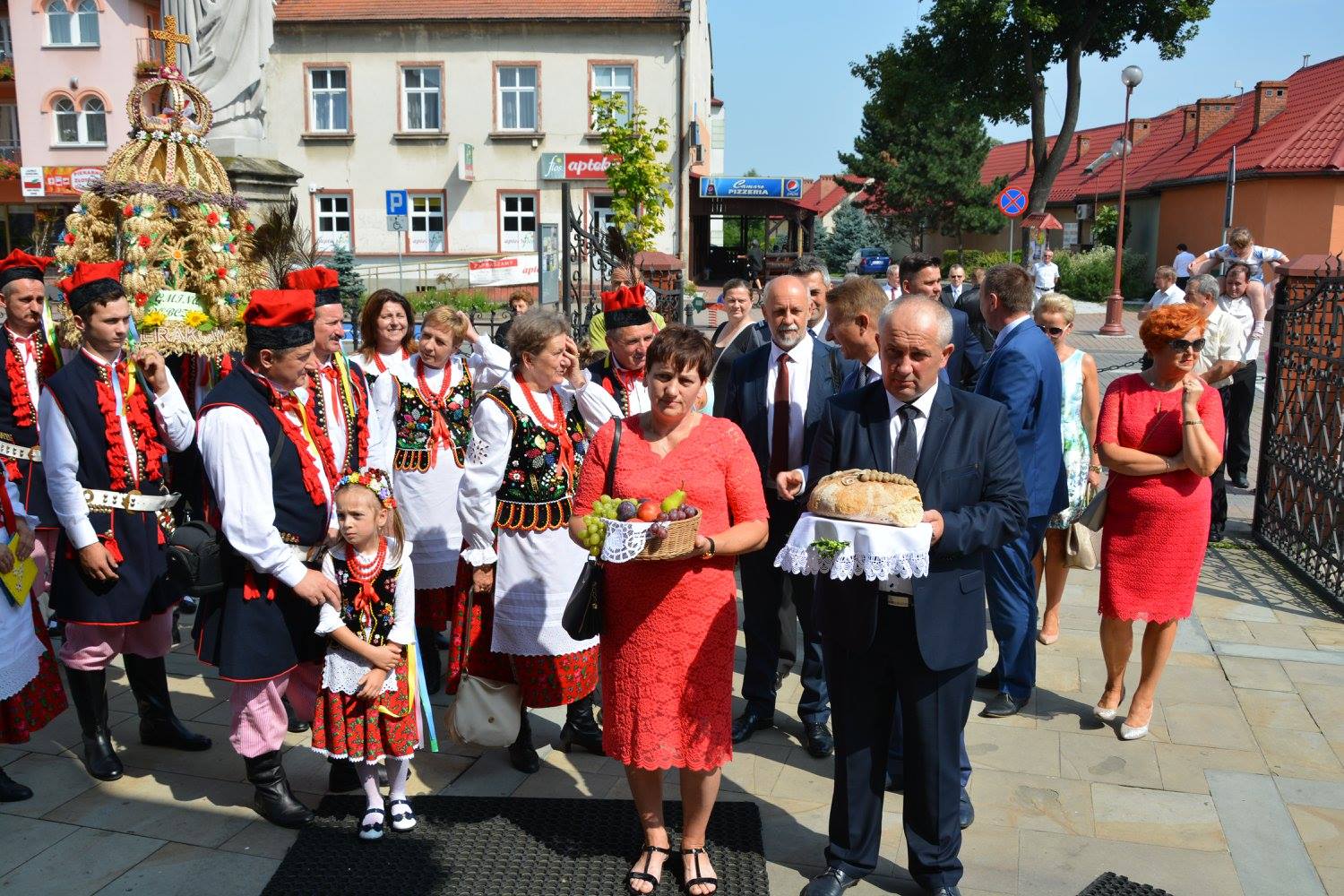 grupy dożynkowe niosą w darze wieńce dożynkowe. Z przodu stoją starostowie dożynek niosący chleb i winogrona