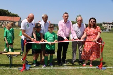 Sport, emocje i pomoc – wyjątkowa niedziela w Stadnikach Na boisku sportowym sześć osób, w tym przedstawiciele samorządu, lokalnej społeczności i młodzi piłkarze, wspólnie przecina biało-czerwoną wstęgę podczas uroczystego otwarcia zmodernizowanego obiektu. W tle stoją dzieci w zielonych strojach piłkarskich oraz widoczne są zabudowania i murawa boiska. Słoneczna pogoda podkreśla uroczysty charakter wydarzenia
