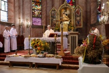 ksiądz odprawiający mszę świętą z boku służba liturgiczna, przed ołtarzem chleby i kołacze ofiarowane przez rolników po bokach ułożone sa wieńce dożynkowe 
