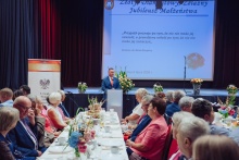 Na pierwszym planie przy długich, nakrytych stołach siedzą uczestnicy uroczystości Jubileuszu Złotych, Diamentowych i Żelaznych Godów. W centralnej części sali przy mównicy przemawia burmistrz Tomasz Suś. W tle widoczna jest scena z dekoracjami kwiatowymi oraz ekran z napisem „Złoty, Diamentowy, Żelazny Jubileusz Małżeństwa” i cytatem Antoine'a de Saint-Exupéry'ego. Na stołach ustawiono kwiatowe kompozycje i poczęstunek, a całość tworzy uroczystą, odświętną atmosferę wydarzenia