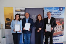 Na zdjęciu cztery osoby stoją podczas uroczystego spotkania - od lewej: przewodnciząca rady miejskiej, wojewoda, kierownik ŚDS oraz burmistrz. Dwie osoby trzymają dyplomy z podziękowaniami. W tle widoczne są roll-upy promocyjne oraz baner Środowiskowego Domu Samopomocy w Dobczycach. Uczestnicy są ubrani w stroje formalne i pozują do wspólnego zdjęcia.