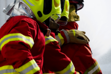 Dwóch strażaków w czerwonych ubraniach ochronnych i kaskach trzyma wąż gaśniczy podczas akcji, w tle widoczny dym.