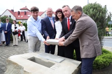 Pięć osób uczestniczy w uroczystości wmurowania kapsuły czasu. Wspólnie trzymają metalowy pojemnik i umieszczają go w specjalnie przygotowanym zagłębieniu w kamiennym postumencie. W tle widoczni są pozostali uczestnicy wydarzenia oraz budynek i zieleń otaczająca miejsce ceremonii. Zdjęcie wykonano na zewnątrz podczas oficjalnych obchodów 120-lecia Szkoły Podstawowej w Kornatce