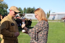 Wręczenie pucharu podczas zawodów sportowo-pożarniczych – druh w mundurze odbiera trofeum od przedstawicielki organizatorów. W tle uczestnicy zawodów i boisko sportowe.