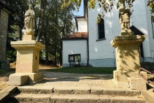 dwie kamienne gigury z ostaciami świętych stojące u szczytu piaskowcowych schodów po obu ich stronach, w tle biała ściana kościoła