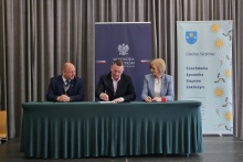 Trzy osoby siedzą przy stole podczas oficjalnego podpisania umowy. W centrum mężczyzna składa podpis na dokumencie, obok niego siedzą kobieta i mężczyzna obserwujący uroczystość. W tle widoczne są banery – jeden z godłem i napisem „Wojewoda Małopolski”, drugi z nazwą „Gmina Siepraw” oraz listą miejscowości.