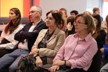 Uczestnicy konferencji siedzący na sali, skupieni na wystąpieniach – na pierwszym planie trzy osoby w średnim wieku, słuchające prelekcji, w tle widoczna grupa pozostałych uczestników wydarzenia.