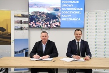 przy stole na siedzą burmistrz Gminy i maista Dobczyce Tomasz Suś z przedstawicielem firmy Vector Software sp. z o.o, pozują do zdjęcia podczas pospisywania umowy, w tle zdjęcie Dobczyc z lotu ptaka z widokiem na jezioro oraz grafiki z logiem Dobczyc