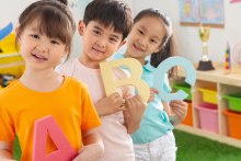 trójka dzieci stoi w sali przedszkolnej, każde z nich trzyma w rękach inną literę alfabetu