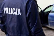 Policjant stojący przy radiowozie, na kurtce widoczny napis „POLICJA"