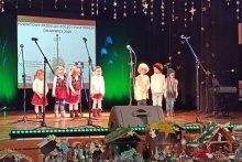 na scenie w rzędzie stoją dzieci przed nimi rozmieszczone są miktofony w tle wyświetlany jest baner o treści Powiatowy Przegląd Kolęd i Pastorałek, wokół świąteczne dekoracje 