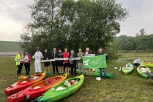 Grupa ludzi stojąca na tle zielonego drzewa. Każdy z nich trzyma napisy. Przed nimi leżą różnego koloru kajaki 