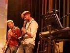 Dwóch mężczyzn grających na gitarach podczas koncertu. Mężczyźni są skupieni.