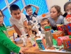 w sali dzieci siedzą a niektóre stoją przy długim prostokątnym stole na stole ustawiona jest wykonana makieta ekologicznego miastai wykonują prace plastyczne 