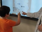 chłopiec stoi przed interaktywną tablicą na której widnieje mapa Polski, wskazuje coś na mapie, widać też inną dłoń która pokazuje punkt na mapie