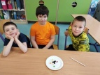 Trzech chłopców siedzi przy szkolnej ławce, na ławce leży talerzyk tekturowy a na nim zbudowany ze słodyczy model atomu 