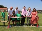 Sport, emocje i pomoc – wyjątkowa niedziela w Stadnikach Na boisku sportowym sześć osób, w tym przedstawiciele samorządu, lokalnej społeczności i młodzi piłkarze, wspólnie przecina biało-czerwoną wstęgę podczas uroczystego otwarcia zmodernizowanego obiektu. W tle stoją dzieci w zielonych strojach piłkarskich oraz widoczne są zabudowania i murawa boiska. Słoneczna pogoda podkreśla uroczysty charakter wydarzenia