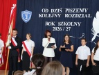 Uczniowie i nauczyciele podczas uroczystości rozpoczęcia roku szkolnego 2026/2027. W tle napis „Od dziś piszemy kolejny rozdział” i szkolny sztandar.