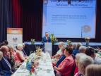 Złote Gody w Gminie Dobczyce – uhonorowano Jubilatów, którzy swoje święto obchodzili w 2025 roku Na pierwszym planie przy długich, nakrytych stołach siedzą uczestnicy uroczystości Jubileuszu Złotych, Diamentowych i Żelaznych Godów. W centralnej części sali przy mównicy przemawia burmistrz Tomasz Suś. W tle widoczna jest scena z dekoracjami kwiatowymi oraz ekran z napisem „Złoty, Diamentowy, Żelazny Jubileusz Małżeństwa” i cytatem Antoine'a de Saint-Exupéry'ego. Na stołach ustawiono kwiatowe kompozycje i poczęstunek, a całość tworzy uroczystą, odświętną atmosferę wydarzenia