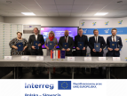 Siedem osób w strojach formalnych stoi za stołem konferencyjnym i trzyma niebieskie teczki z podpisanymi dokumentami. Na stole znajdują się wizytówki, długopisy oraz małe flagi Unii Europejskiej i państw partnerskich. W tle widoczny jest ekran z informacją o podpisaniu umowy o kontynuacji współpracy oraz roll-upy z logo Gminy Dobczyce i programu Interreg Polska–Słowacja.