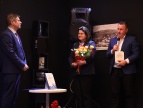 Na zdjęciu widać trzy osoby podczas spotkania promocyjnego książki w bibliotece. Po lewej stronie stoi mężczyzna w garniturze - autor, zwrócony w stronę pozostałych uczestników, słuchający wypowiedzi. Po prawej stronie mężczyzna w marynarce – burmistrz, trzyma mikrofon i książkę pt. „Miasto Dobczyce i jego wiejskie otoczenie w latach 1918–1939”, przemawiając do zgromadzonych. Pośrodku stoi kobieta w ciemnym stroju - przewodnicząca, trzymająca bukiet kwiatów. Przed nimi znajduje się niewielki stolik z książk