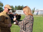 Wręczenie pucharu podczas zawodów sportowo-pożarniczych – druh w mundurze odbiera trofeum od przedstawicielki organizatorów. W tle uczestnicy zawodów i boisko sportowe.