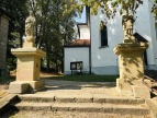 dwie kamienne gigury z ostaciami świętych stojące u szczytu piaskowcowych schodów po obu ich stronach, w tle biała ściana kościoła