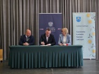 Trzy osoby siedzą przy stole podczas oficjalnego podpisania umowy. W centrum mężczyzna składa podpis na dokumencie, obok niego siedzą kobieta i mężczyzna obserwujący uroczystość. W tle widoczne są banery – jeden z godłem i napisem „Wojewoda Małopolski”, drugi z nazwą „Gmina Siepraw” oraz listą miejscowości.