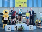Pan Burmistrz stojący na podium z uczestnikami V Rodzinnego mini festiwalu "o złotą kózkę". Maskotka stojąca obok uczestników przebrana jest za słońce w żółtym kolorze 