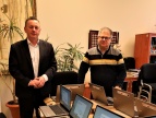 Burmistrz Tomasz Suś oraz dyrektor Szkoły Podstawowej nr 2 w Dobczycach, Aleksander Płoskonka stoją w pomieszczeniu biurowym obok stołu, na którym ustawiono kilka laptopów umieszczonych na kartonowych pudełkach. Na laptopach widoczne są naklejki z symbolami Unii Europejskiej i Polski. W tle znajdują się rośliny doniczkowe, biurowe meble oraz obrazy na ścianach.