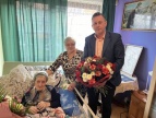 starsza kobieta leżąca na łóżku a tu obok niej mężczyzna w garniturze z kwiatami w ręku 