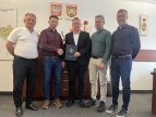 Rozbudowa sieci wodociągowej - blisko kilometr wodociągu powstanie na ul. Grabowej Pięciu mężczyzn stoi w biurze urzędu. Dwóch mężczyzn pośrodku ściska sobie dłonie - burmistrz oraz przedstawiciel wykonawcy, a jeden z nich trzyma granatową teczkę z herbem i napisem „Gmina i Miasto Dobczyce”. W tle widoczne są godła i symbole urzędowe zawieszone na ścianie oraz meble biurowe. Zdjęcie dokumentuje podpisanie umowy.