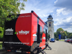 Czerwony mobilny punkt programu Czyste Powietrze zaparkowany przy ulicy, w tle budynek z wieżą zegarową.