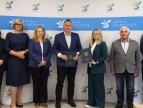 Rusza kolejny etap przebudowy drogi gminnej w Nowej Wsi i Sierakowie Siedem osób pozuje do pamiątkowego zdjęcia na tle ścianki promocyjnej Gminy Dobczyce. W centrum stoją przedstawiciele samorządu oraz wykonawcy robót, trzymając podpisane dokumenty umowy w granatowych teczkach z herbem gminy. Pozostałe osoby stoją po obu stronach grupy. Wszyscy ubrani są w stroje biznesowe lub półformalne. Zdjęcie wykonano w pomieszczeniu urzędu po zakończeniu oficjalnego podpisania dokumentów.