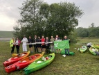 Grupa ludzi stojąca na tle zielonego drzewa. Każdy z nich trzyma napisy. Przed nimi leżą różnego koloru kajaki 