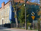 Duży budynek z niebieską folia po jednej stronie. Przed budynkiem widnieje pomarańczowy znak i biały z trójkątem w środku