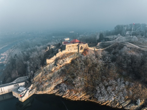 Zamek wzniesiony na skalistym wzgórzu nad rzeką, widziany z lotu ptaka w zimowej scenerii. Kamienne mury i czerwony dach kontrastują z oszronionymi drzewami i mgłą unoszącą się nad okolicą. Po prawej stronie widoczny jest maszt z polską flagą, a w tle zarys zabudowy miejscowości.